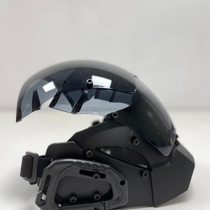 May include: Black tactical helmet with a clear, curved visor. The helmet features a chin guard, adjustable strap, and multiple attachment points. The visor is slightly tinted, and the helmet has a matte finish.