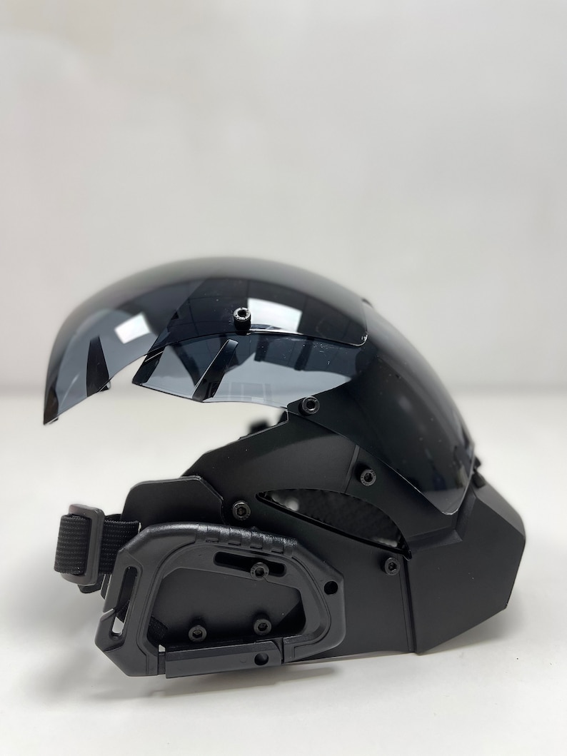 May include: Black tactical helmet with a clear, curved visor. The helmet features a chin guard, adjustable strap, and multiple attachment points. The visor is slightly tinted, and the helmet has a matte finish.