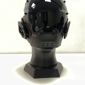 May include: A glossy black futuristic helmet with a visor and integrated ear pieces. The helmet is mounted on a black display stand. The design features a mouth guard with ventilation holes, suggesting a focus on both aesthetics and functionality.