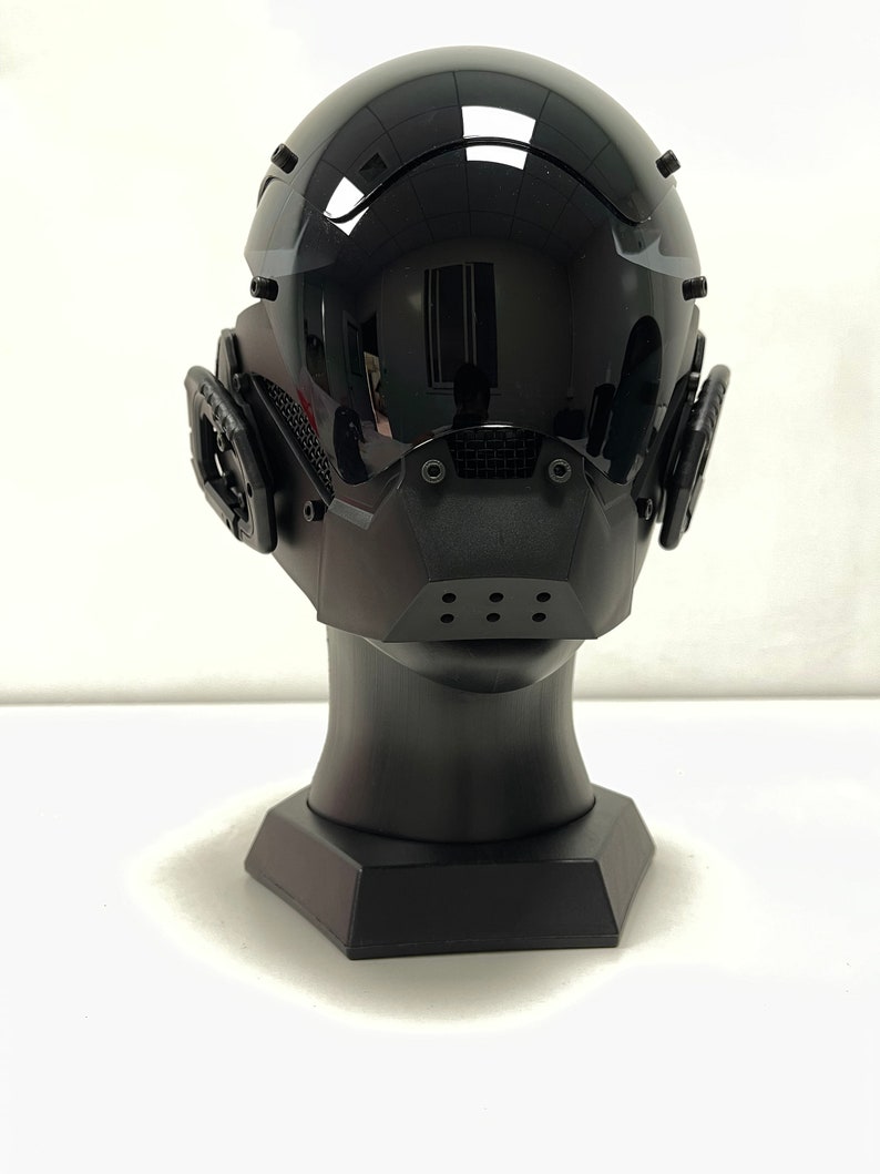 May include: A glossy black futuristic helmet with a visor and integrated ear pieces. The helmet is mounted on a black display stand. The design features a mouth guard with ventilation holes, suggesting a focus on both aesthetics and functionality.