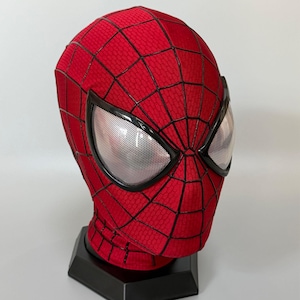 May include: A red fabric mask featuring a black web pattern across the surface. The mask includes two large, almond-shaped eye lenses with a silver, mesh-like texture framed in black, resting on a black hexagonal display stand.