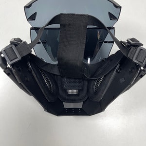 May include: Black tactical mask with a wraparound visor and adjustable straps. The mask features a mesh section over the mouth and nose area, and padded interior for comfort. The visor is tinted and provides eye protection.