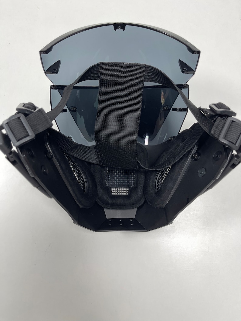May include: Black tactical mask with a wraparound visor and adjustable straps. The mask features a mesh section over the mouth and nose area, and padded interior for comfort. The visor is tinted and provides eye protection.