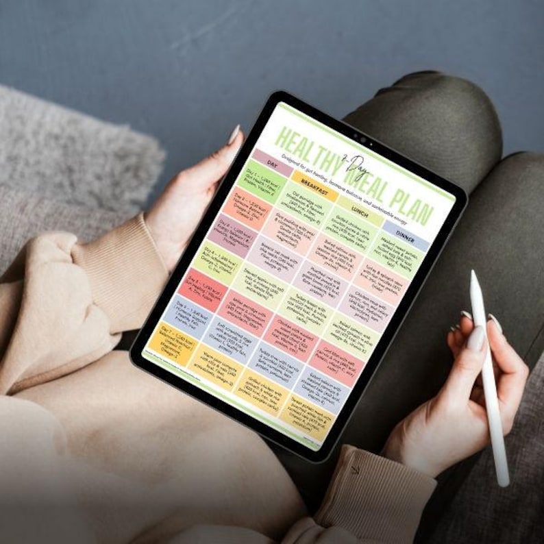 May include: A digital tablet displaying a "Healthy 7-Day Meal Plan" with colorful boxes for each day's meals. The tablet is held by a person, with a white stylus resting in the other hand. The plan is designed for getting, hormone balance, and sustainable energy.