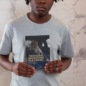 May include: A person holding a book titled "The Digital Marketers Playbook" with a woman on the cover talking on a phone in a car. The book cover is black with gold text.