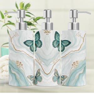 May include: Three decorative soap dispensers with silver pumps. The dispensers feature a white background with a teal and gold marble design and teal butterfly illustrations. The dispensers are cylindrical in shape.