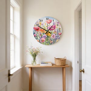 May include: A round wall clock featuring a colorful watercolor illustration of various wildflowers including poppies and lavender. The clock face has black numbers from 1 to 12 and thin black hour, minute, and second hands centered on the floral design.