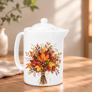 May include: A white ceramic teapot with a lid and a handle, featuring a colorful printed bouquet of autumn leaves, berries, and twigs tied with a bow. The design displays shades of orange, red, yellow, and brown on the front surface.