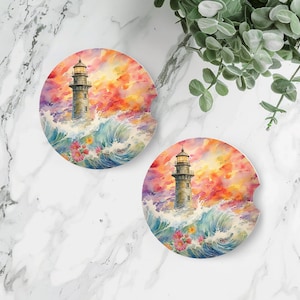 May include: Two round coasters with a watercolor design of a lighthouse in the ocean with a sunset sky. The lighthouse is white with a black roof and a red light at the top. The ocean is blue with white waves. The sky is orange, pink, and purple. There are flowers in the water around the lighthouse.