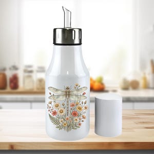 May include: White oil dispenser with a stainless steel spout and lid. The dispenser features a dragonfly and floral design in shades of beige, orange, and yellow. A white cylindrical stopper sits beside the bottle.