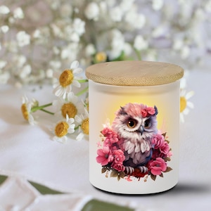 May include: A white candle jar with a bamboo lid features a watercolor illustration of an owl surrounded by pink flowers. The owl has pink, purple, and white feathers and large brown eyes. The background is softly lit.