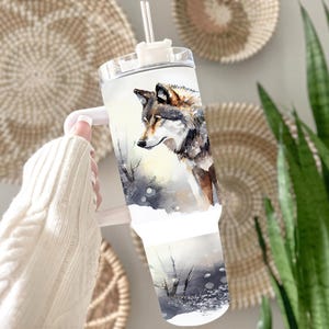 A person's hand holding a tumbler with a printed image of a wolf on it, set against a backdrop of woven baskets and plants.