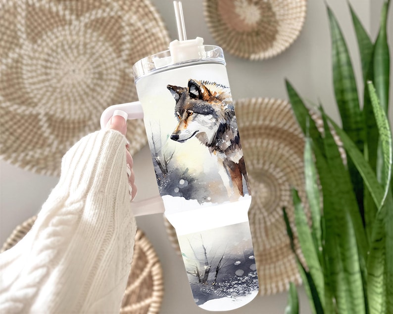A person's hand holding a tumbler with a printed image of a wolf on it, set against a backdrop of woven baskets and plants.