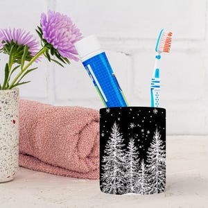 May include: A black and white toothbrush holder with a winter forest design, holding a tube of toothpaste and a toothbrush. A pink towel and a vase with purple flowers are in the background.