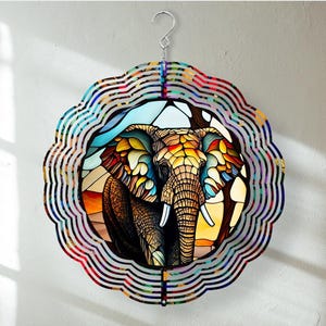 May include: A colorful wind spinner featuring an elephant design. The elephant is depicted in a stained-glass style with vibrant hues of orange, yellow, blue, and brown. The spinner has a scalloped edge and a silver hook for hanging.