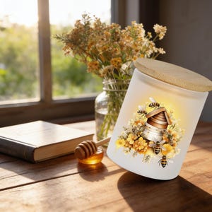May include: A frosted glass jar with a light wood lid, featuring a colorful graphic of a wooden beehive surrounded by yellow flowers and several bees. The design includes honey-like drips and a warm, glowing yellow background behind the hive.