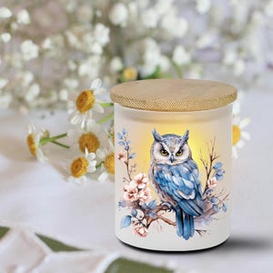 May include: A white candle holder with a bamboo lid features a watercolor owl design. The owl is blue and gray, perched on a branch with pink and blue floral accents. The candle holder is surrounded by white flowers.