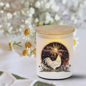 May include: A white candle jar with a wooden lid. The jar features a circular illustration of a rooster, sun, and flowers. The background includes white flowers.