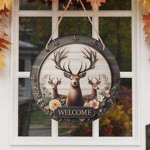Op de afbeelding: Een ronde houten welkomstbord met een hertenfamilie met grote geweien en bloemaccenten. Het bord heeft een donkere houten rand en een lichte houten achtergrond met het woord "WELCOME" onderaan. Het hangt aan een witte deur.
