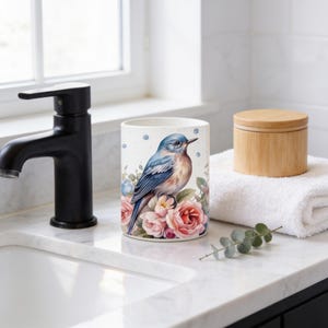 May include: A white cylindrical ceramic tumbler featuring a detailed illustration of a blue bird perched among soft pink and peach-colored roses. The design includes small blue circular accents scattered around the bird and floral arrangement.