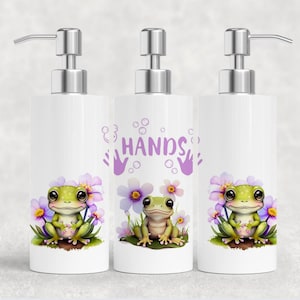 May include: Three white ceramic soap dispensers with silver pumps. Each dispenser features a cartoon frog design with purple flowers and the word "HANDS" in purple. The frogs are green with large eyes.