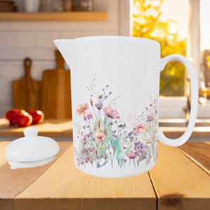 May include: White ceramic pitcher with a floral design featuring pink, purple, and green wildflowers. The pitcher has a handle and a spout, and a matching lid is next to it. The pitcher is on a wooden surface.