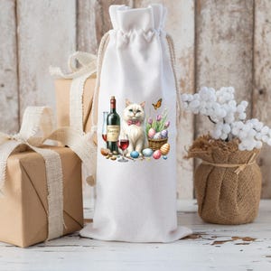 May include: White wine bottle gift bag with a white cat wearing a bow tie and holding a glass of wine. The cat is surrounded by Easter eggs and a butterfly. The bag is made of natural fabric.