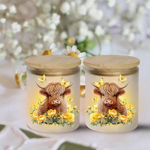 May include: Two glass jars with wooden lids. Each jar features a Highland cow illustration with yellow butterflies and flowers. The jars are cylindrical and have a frosted appearance, with a soft, warm glow.