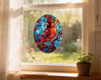 May include: An oval stained glass suncatcher featuring a vibrant red cardinal bird perched on a brown branch. The background consists of blue geometric glass pieces and small red and yellow flowers. It hangs from a clear suction cup hook.