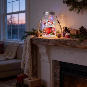 May include: A frosted glass mason jar lantern featuring a graphic of four snowmen in a red mug, illuminated by internal multi-colored fairy lights. The jar has a silver metal lid and a thick, natural-colored rope handle attached to the sides.
