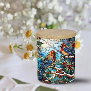 May include: A cylindrical container with a stained-glass design featuring two birds perched on a branch with red berries. The lid is made of light-colored wood. The background includes white flowers.