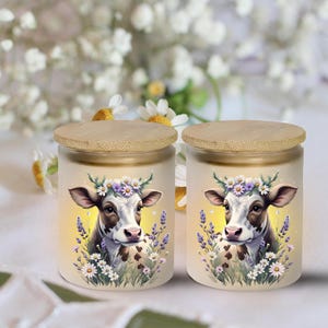May include: Two glass jars with bamboo lids. Each jar features a cow illustration wearing a floral crown, surrounded by lavender and daisies. The background is a soft yellow gradient.