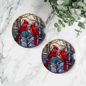 May include: Two red cardinal birds perched on branches in a wintery forest scene. The birds are surrounded by blue and white stained glass-like details. The image is a round coaster.