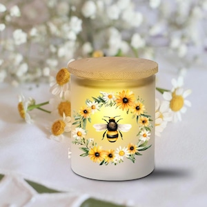 May include: A white candle holder with a wooden lid, featuring a bee and floral design. The candle holder is decorated with a wreath of yellow sunflowers, white daisies, and green leaves. The bee is centered in the design, with a yellow and black striped body.