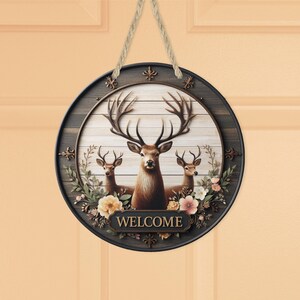 Op de afbeelding: Ronde houten welkomstbord met een hertenfamilie met grote geweien en bloemaccenten. Het bord heeft een donkerbruine rand en een lichtbruine achtergrond met het woord "WELCOME" onderaan. Een natuurlijk touw wordt gebruikt om op te hangen.