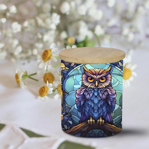 May include: A cylindrical container with a bamboo lid. The container features a stained-glass style owl design in shades of blue, purple, and yellow. The owl is perched on a branch against a blue background. Daisies are in the background.