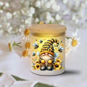May include: A white jar with a wooden lid featuring a whimsical gnome design. The gnome wears a yellow and black striped hat and is surrounded by bees and sunflowers. The jar is approximately 4 inches tall.