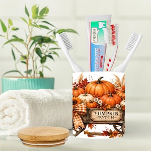 May include: A cylindrical white toothbrush holder featuring a colorful autumn graphic of pumpkins, fall leaves, and a wooden cart with the text PUMPKIN PATCH. The holder contains two white toothbrushes and a tube of toothpaste with red and blue accents.