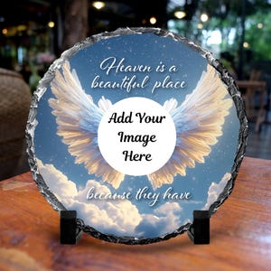 May include: A round, natural stone plaque with a sky-blue background, white clouds, and angel wings. The text "Heaven is a beautiful place" and "Add Your Image Here" are displayed. The plaque is supported by a black stand.
