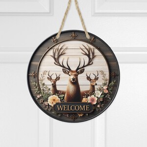 Op de afbeelding: Ronde houten welkomstbord met drie herten met grote geweien en bloemaccenten. Het bord heeft een donkere rand en een lichte houtnerf achtergrond. Het woord "WELCOME" wordt weergegeven op een rechthoekige plaat.