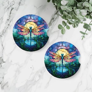 May include: Two round coasters with a stained glass design featuring a dragonfly with pink and yellow wings against a blue and purple background. The dragonfly is centered on each coaster and is surrounded by a circular pattern of blue, purple, and yellow.