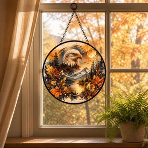 May include: A circular hanging suncatcher featuring a detailed illustration of a bald eagle head, snow-capped mountains, pine trees, and a lake, all framed by vibrant orange and yellow autumn leaves, suspended by a metal chain against a window.