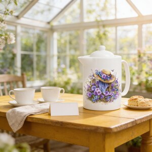 May include: A white ceramic teapot featuring a colorful illustration of a straw hat adorned with purple and blue flowers. The teapot is placed on a wooden table alongside two white teacups, a folded beige napkin, and a small blank white card.