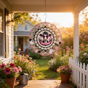 May include: A circular metal wind spinner featuring a central graphic of a cow wearing pink sunglasses and a pink ribbon. The text "SAVE THE UDDERS" is printed below the cow. The spinner has a scalloped edge with alternating black and pink patterns.