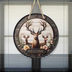 Op de afbeelding: Ronde houten welkomstbord met drie herten en bloemendetails. Het bord heeft een donkerbruine rand en een lichtbruine achtergrond, met het woord "WELCOME" op een gouden banner.