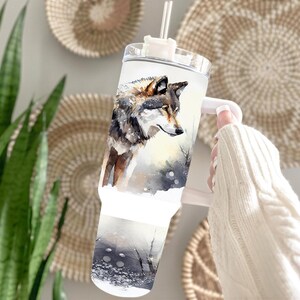 A person's hand holding a clear glass mug with a printed image of a wolf on it, against a backdrop of woven baskets and a potted plant.