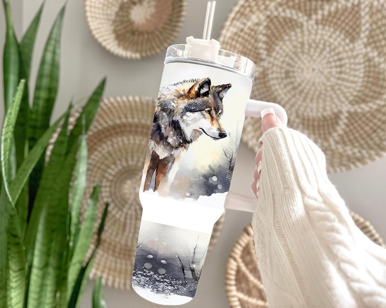 A person's hand holding a clear glass mug with a printed image of a wolf on it, against a backdrop of woven baskets and a potted plant.