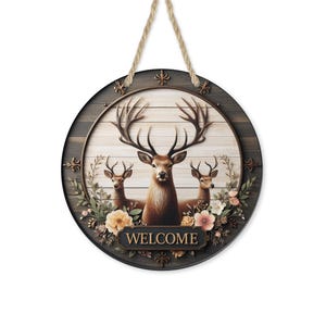 Op de afbeelding: Ronde houten welkomstbord met een centraal beeld van een hert en twee kleinere herten, omgeven door bloemversieringen. Het woord "WELCOME" staat onderaan het bord, dat aan een touw hangt.