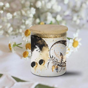 May include: A white candle jar with a gold and black moon and feather design. The jar has a wooden lid and is decorated with gold glitter.