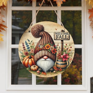 May include: Round wooden welcome sign with a gnome wearing a knit hat and surrounded by pumpkins, flowers, and a barrel. The sign reads "WELCOME FALL" on a wooden sign. The sign is hanging on a white door with fall leaves.